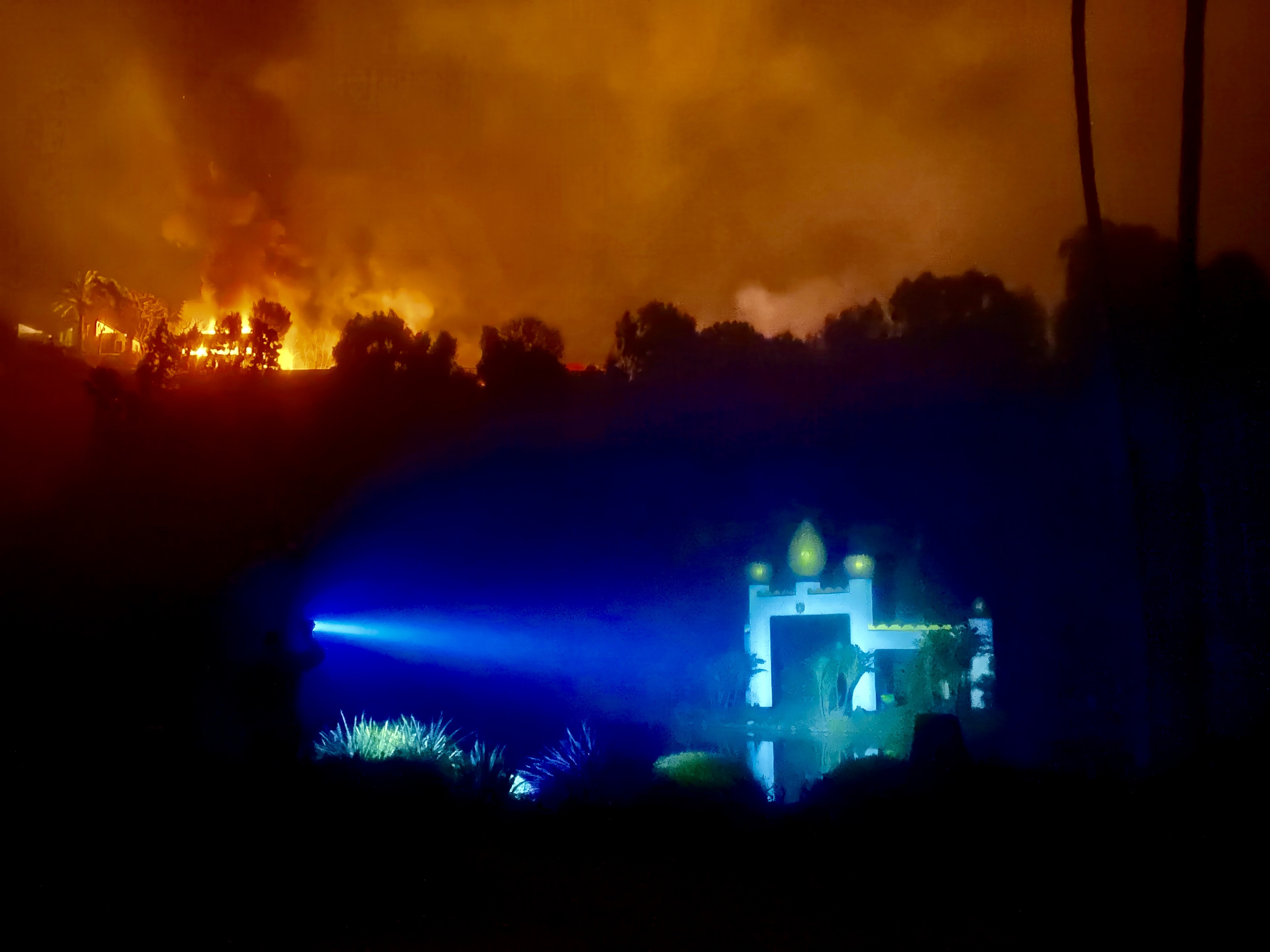 The Lake Shrine's illuminated temple archway stands lit by a single beam as wildfire burns on the ridge beyond.